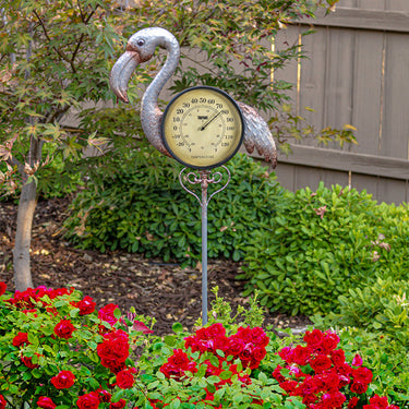 Flamingo Thermometer Garden Stake. Round thermometer as part of a silver flamingo attached to a tall silver garden stake. Shown how pretty it is in a garden with greenery and read flowers.