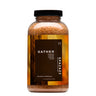 A clear bottle filled with light brown bath salts in the scent "Gather" with notes of pumpkin, nutmeg, orange, and cedar