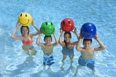  16" Smile Play Beach Balls – 4-Pack - colors are yellow, green, red and blue smiley beach balls. Picture shows children holding one over their heads. 