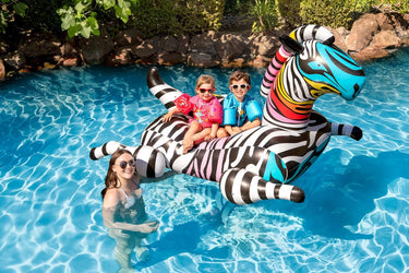 Very large rainbow color zebra pool float. Shown with 2 children riding zany zebra and mom standing next to him in the water.