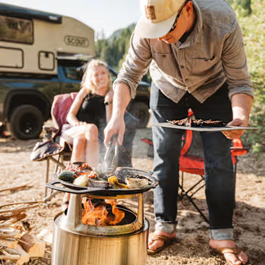 Man grilling food over a campfire with a woman sitting in the background near a camper van.