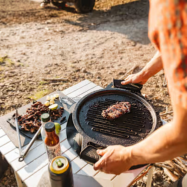 Person grilling outdoors using a portable grill with food and condiments on a wooden table.