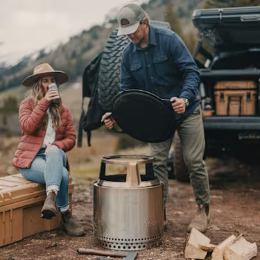 Two people preparing for a camping trip with a portable stove and wood.