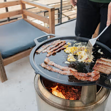 Person cooking bacon and eggs on a cast iron skillet over an open flame on a patio.