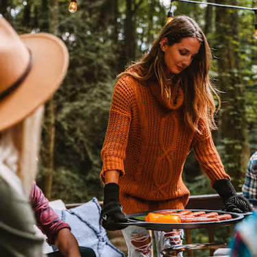 Woman in an orange sweater preparing food on a grill outdoors with trees in the background