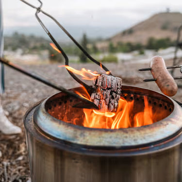Hot dogs being cooked over an open flame in a metal container outdoors.