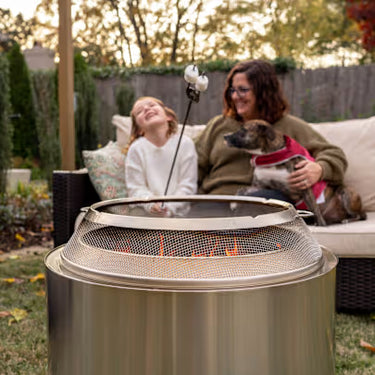 Woman and child sitting on a couch with a dog by a fire pit in a backyard setting.