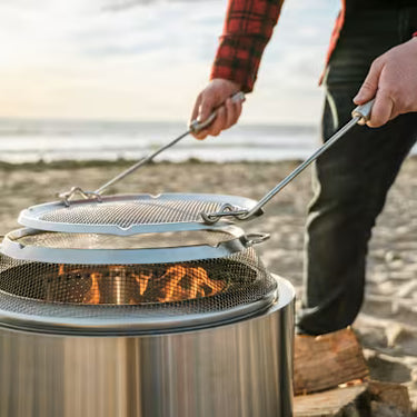 Person using a metal grill over a fire pit on a beach.
