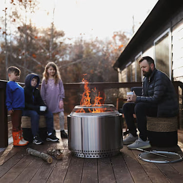 Family sitting around a fire pit on a wooden deck with trees in the background