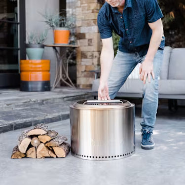Person setting up a stainless steel fire pit with wood logs outdoors.